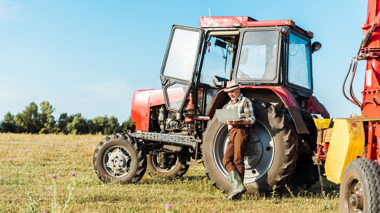 selective focus of bearded farmer in straw hat using laptop near tractor